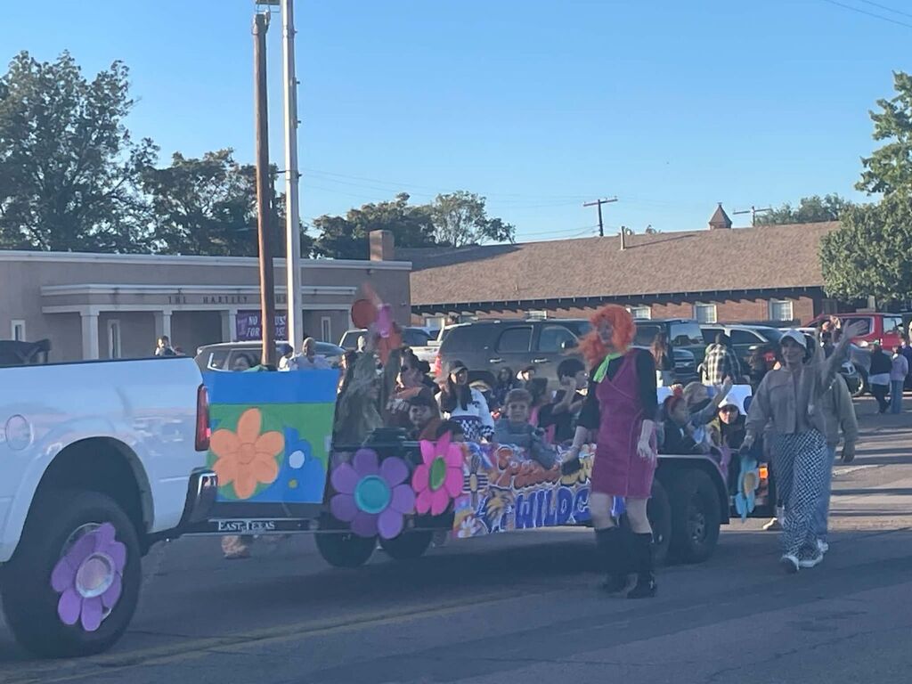 Colorful float in parade with floral designs. Two women walk past a parked white truck. Buildings and trees in the background.