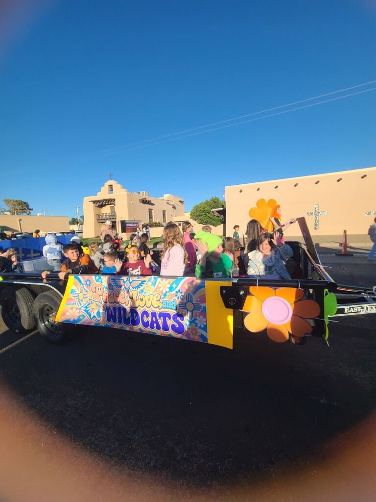 Float with people on it. Banner says "LOVE WILDCATS". Flower design on front. Behind float, buildings and blue sky.