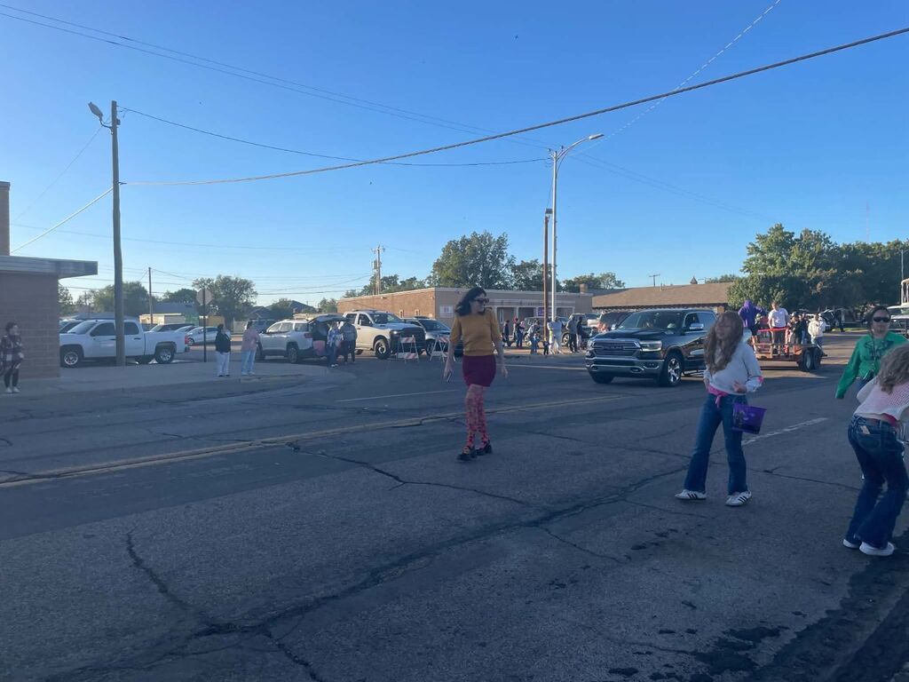 A street with several people walking and standing. Vehicles are parked or driving on the road.