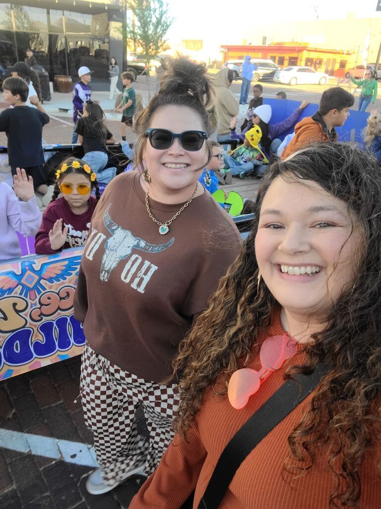 Two women standing and smiling at an outdoor event; one wears sunglasses, the other has goggles. Background includes a crowd and tents.