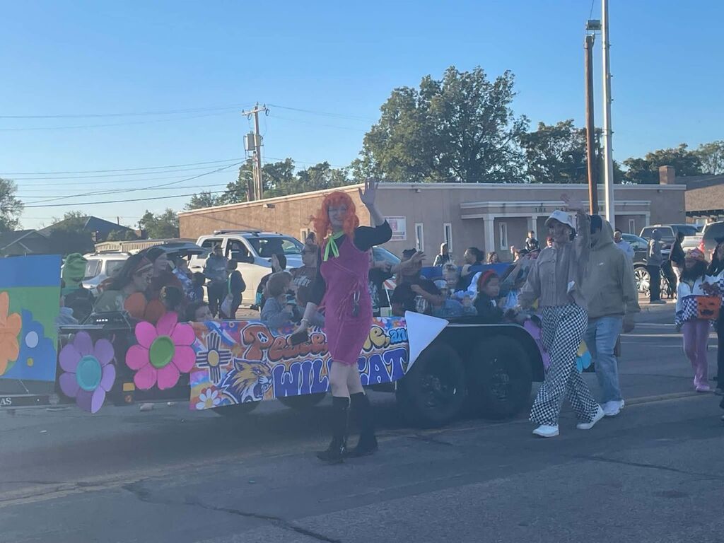 Float with colorful flowers, pumpkin, and Halloween theme. Two people in front, one in costume. Spectators on side.