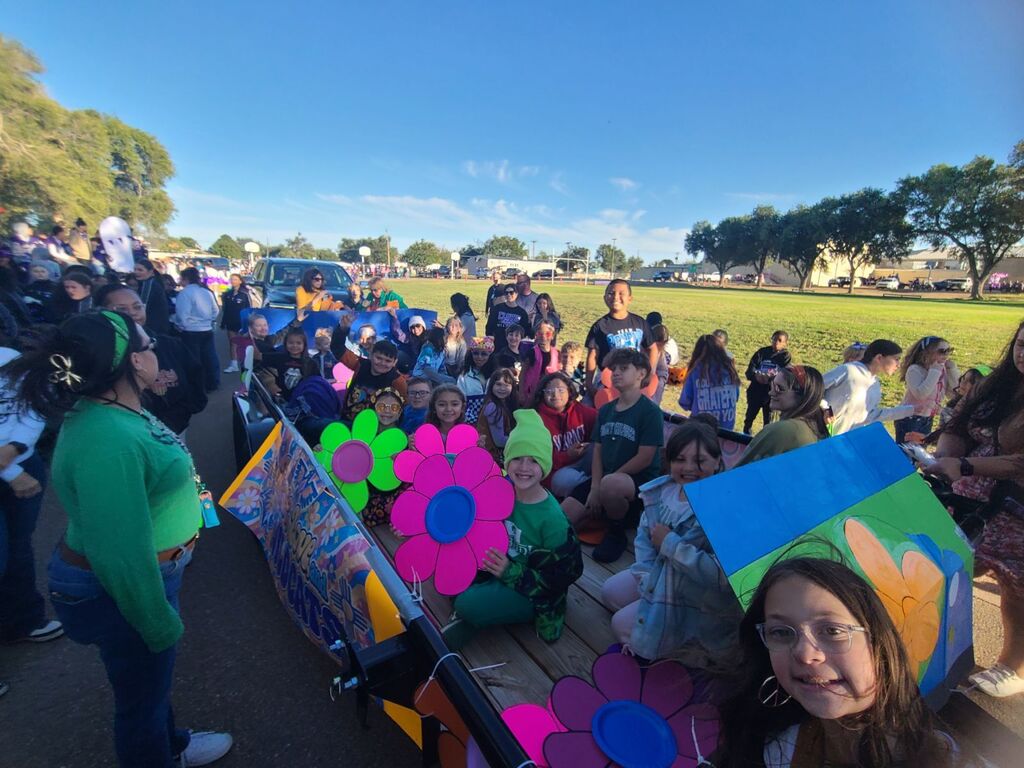 A large group of people stand on a decorated float in a grassy field. Some hold large flowers.