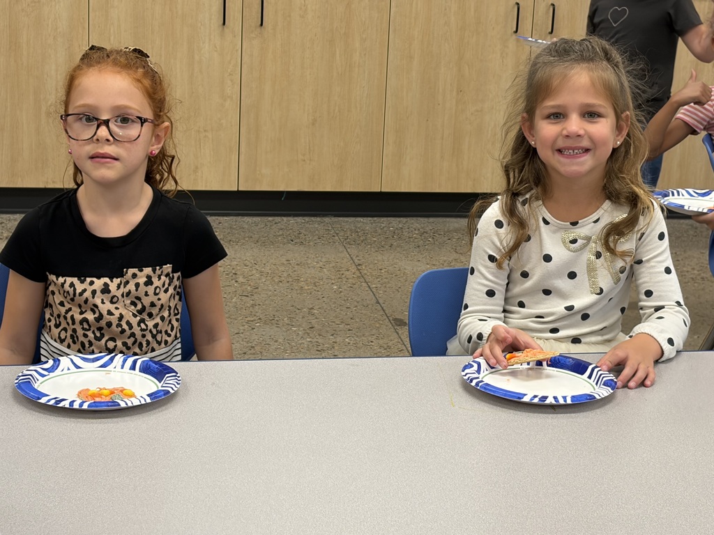 Two young girls seated at a table, smiling; one with glasses, the other holding a sandwich.