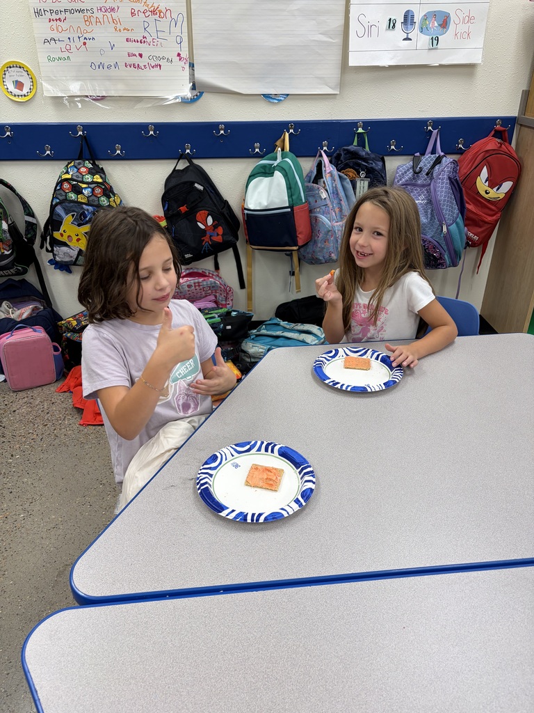 Two girls seated at a table eat a food item on paper plates with blue trim.