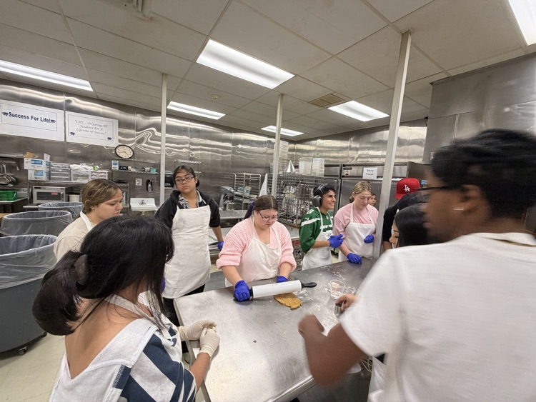 Students and staff in a kitchen. Some are wearing aprons and gloves and are preparing food.