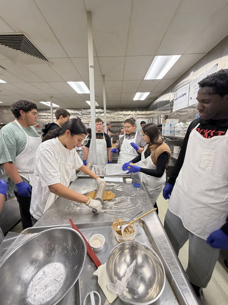 Students wearing white aprons and blue gloves work in a kitchen with rolling pins and mixing bowls.