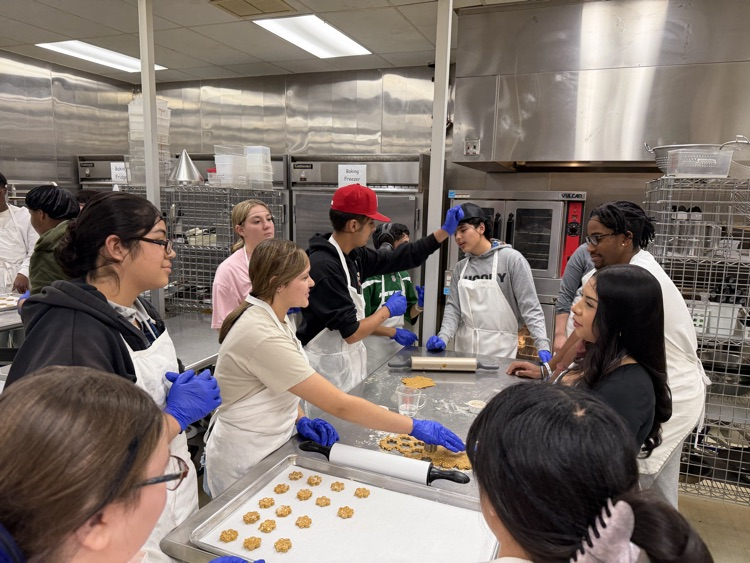 People in aprons and gloves in a kitchen, some standing around and others cutting dough.