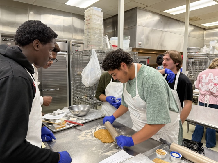 Students in a kitchen with aprons and blue gloves, rolling out dough on a table.