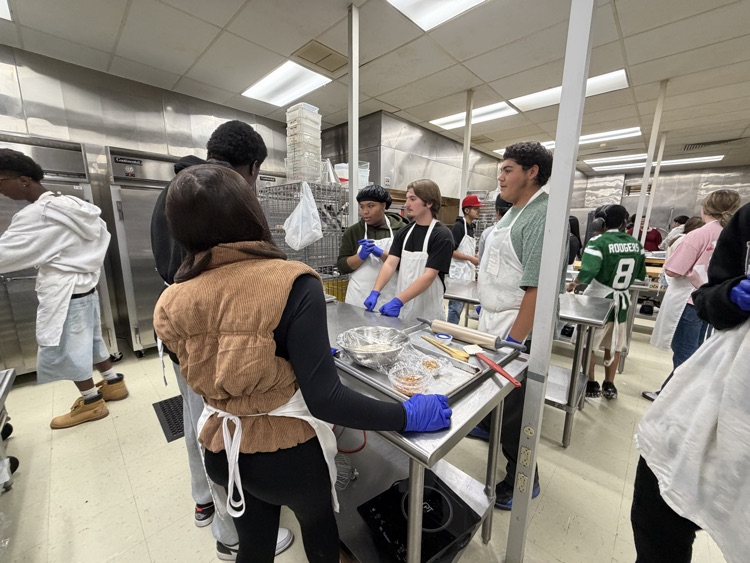 A group of people in aprons and gloves in a kitchen, with two individuals standing at a table.