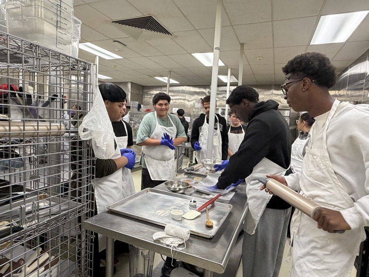 A group of students in white aprons and gloves work at a table in a commercial kitchen.
