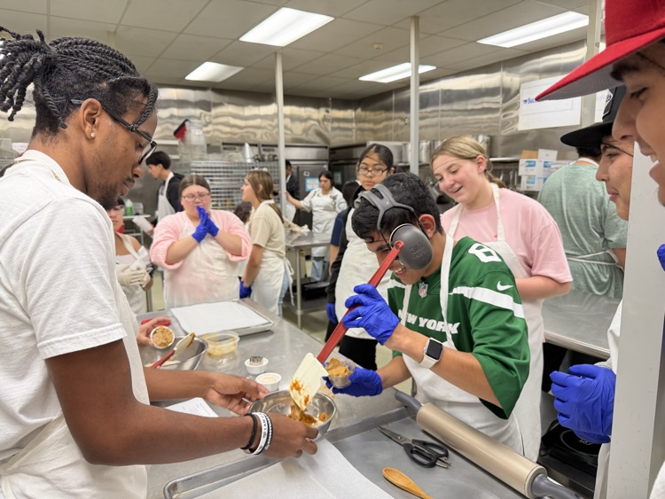 People in a commercial kitchen prepare food. One person stirs a pot while others work with food in bowls.