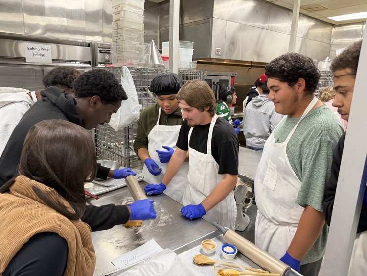 Students in a kitchen with aprons and gloves, preparing food on a counter. Some are rolling dough.