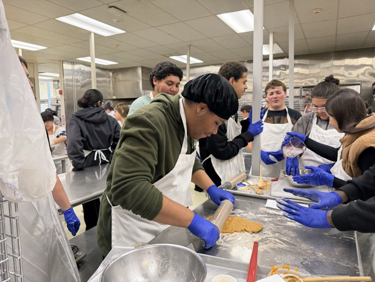A group of students in a kitchen wearing aprons and gloves, preparing food at tables.