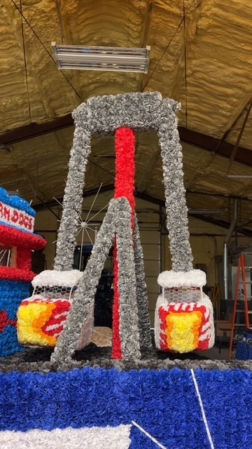 The famous Cole Camp Fair ride "The Bullet" is depicted fully covered in tissue-paper flowers. It has silver arms holding the bullet cages, which are white, with decorative flames.