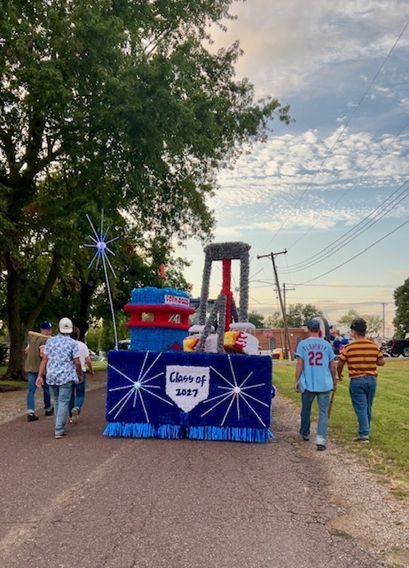 Senior class members are dressed up as The Sandlot characters and walking next to the float as it enters the parade grounds in Cole Camp. The "Class of 2027" is shown on the back of the float on a home plate. 