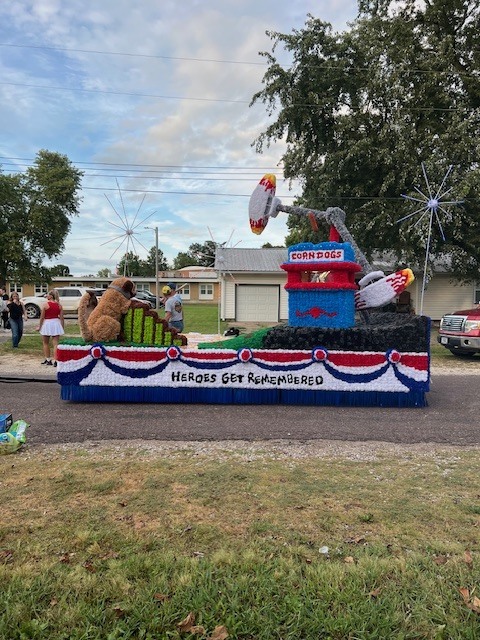 The entire float is pictured, showing the famous dog "Hercules" on the left, jumping over the fence with the baseball in its mouth. The side of the float reads "Heroes get remembered, but legends never die". The Bullet is seen swinging in motion. There is a blue and red corndog stand, mimicking the Craig's Concessions corndog stand at the fair. On the other side, a baseball field is shown. 