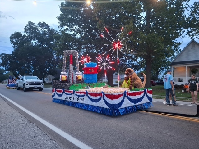 The float is in view depicting the baseball field. The LED fireworks are lit up. You can see the dog jumping over the fence on the right, with the baseball in its mouth. The bullet ride is on the back left, with the corndog stand in blue and red on the right. 