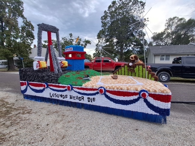 A close photo of the side of the float, reading "legends never die". The baseball field is in plain view, made of sand-colored flowers. It transitions on a slope to outfield grass and then concrete colors as it depicts the carnival scene using the bullet from the Cole Camp Fair and Craig's Concessions corndog stand. 
