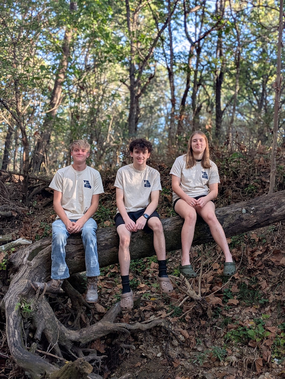 Left to right: Dakota Hagen, Hunter Davis and Mia Fueston sitting on log in wooded area.