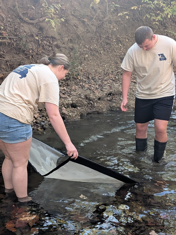 Maddi Root and Chaning Alumbaugh digging for macroinvertebrates standing in creek holding net.