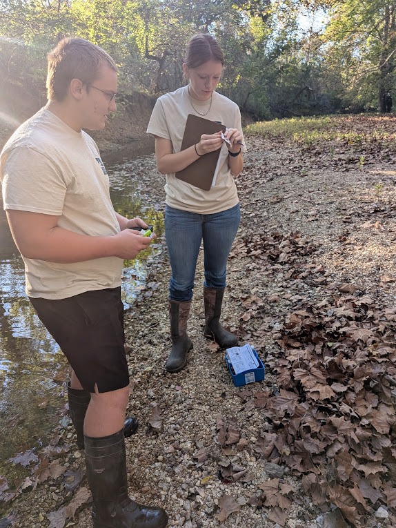 Carson Webb and Leighton Tinney involved in nitrate testing standing on rocky bank of creek in wooded area.