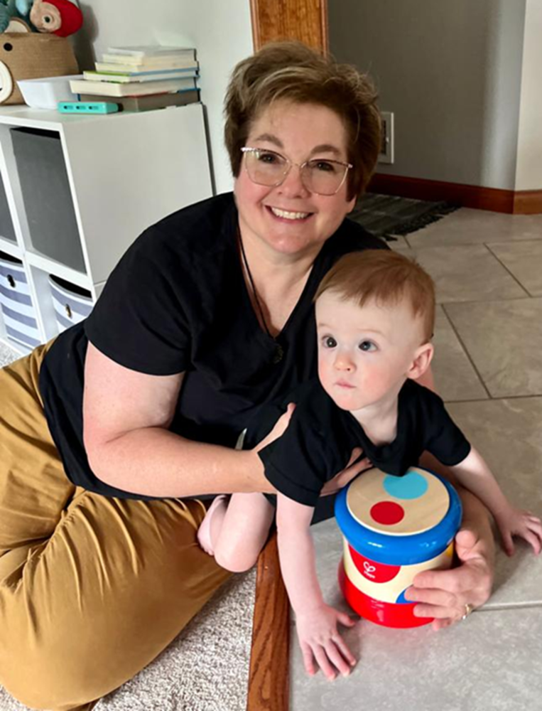 Mrs. Beeson kneels on the floor beside a toddler playing with a colorful toy drum in a cozy home setting. Shelves with books and storage bins are visible in the background.  Congrats Mrs. Beeson on your Retirement!! Thank you for all your years of service here at CC. We will truly miss you! #EagleForever