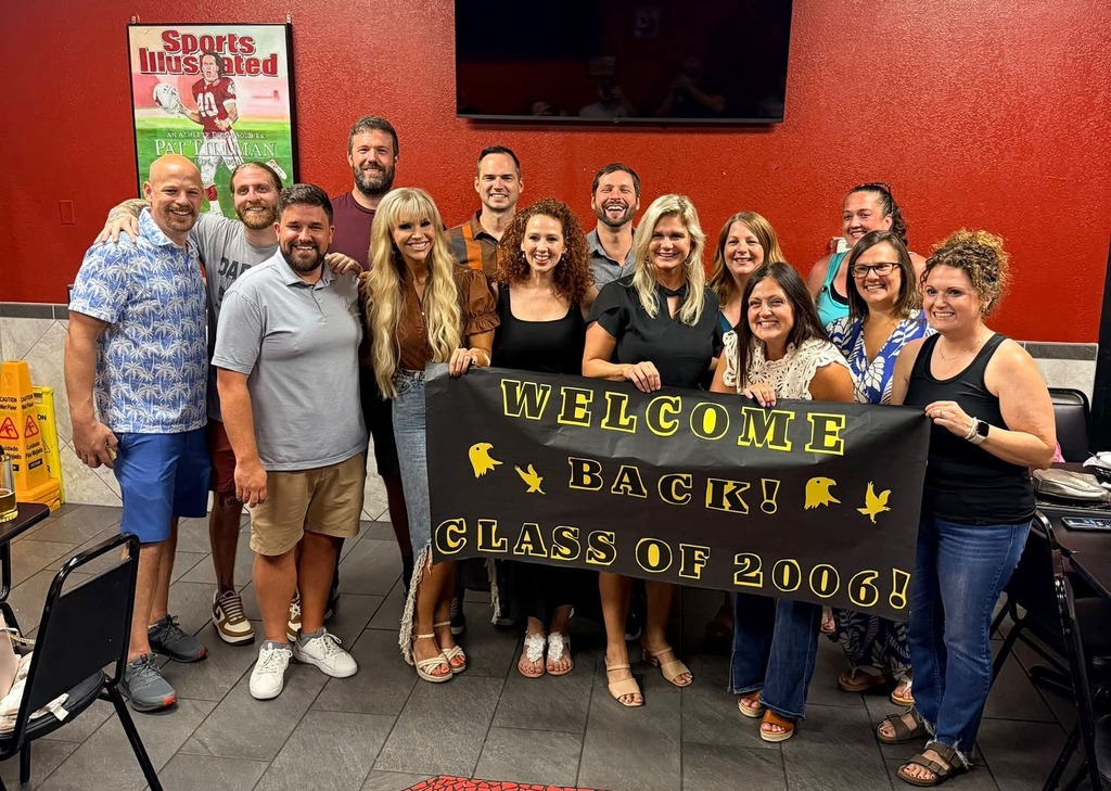 Reunion group photo of adults standing indoors at a casual restaurant, smiling and holding a large black banner with yellow text that reads “WELCOME BACK! CLASS OF 2006!” decorated with small yellow bird graphics, with red walls, tiled floor, chairs, and a “Sports Illustrated” Pat Tillman poster visible in the background.