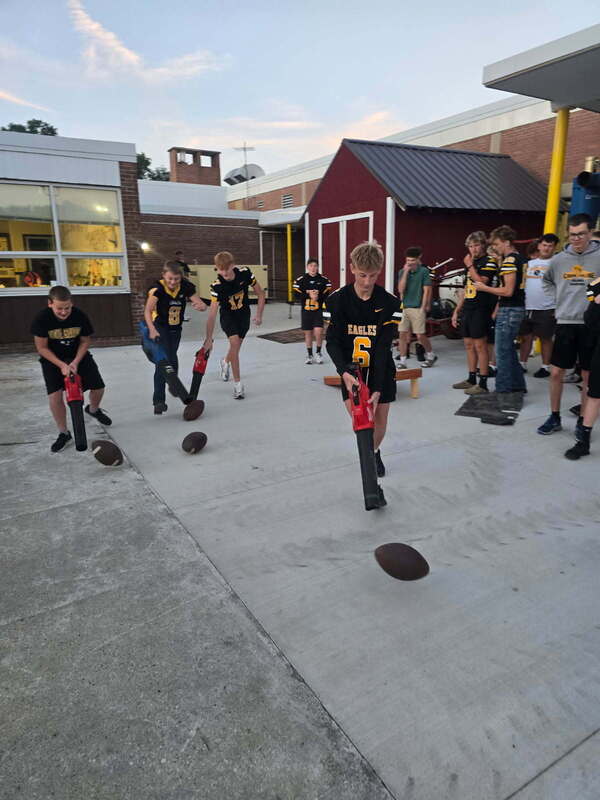 Colonel Crawford students participate in a football race activity outside the school, using leaf blowers to move footballs across the pavement. Several students wear black-and-gold Eagles jerseys while classmates watch and cheer.
