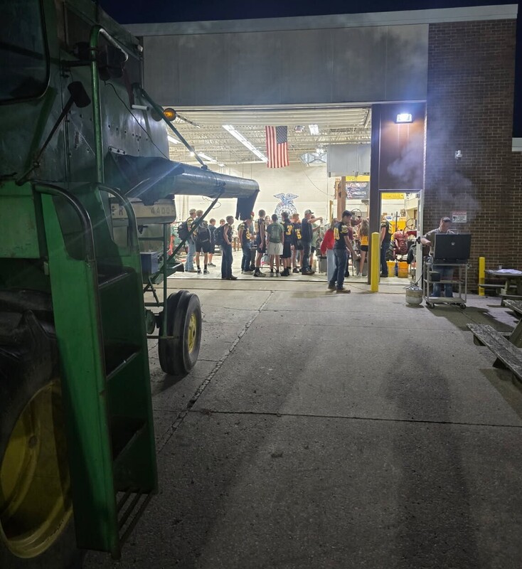Colonel Crawford students gather inside the Ag Shop for an early-morning breakfast event. Several students wear black-and-gold Eagles jerseys, with agricultural equipment, an American flag, and a large green combine visible around the shop.