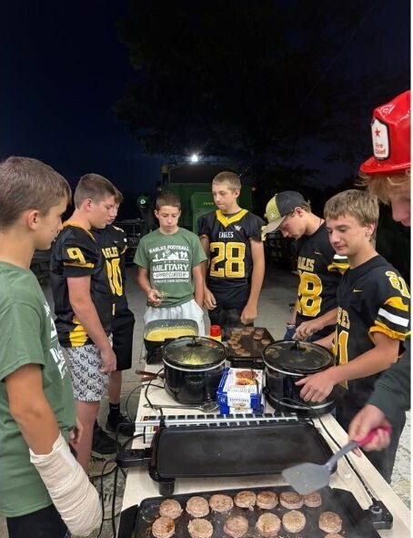 Colonel Crawford students prepare breakfast outdoors before a football game. Several students wearing black-and-gold Eagles football jerseys cook sausage patties and other breakfast foods on electric griddles and slow cookers, while another student wears a red firefighter-style hat.
