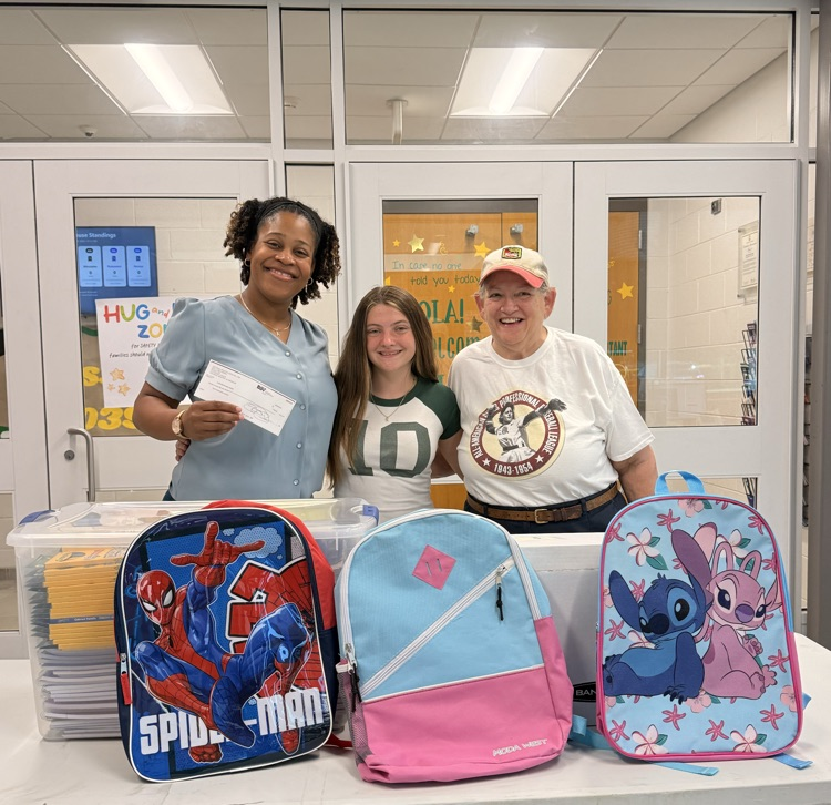 Three people pose behind a table displaying donated school supplies, including backpacks, notebooks, folders, and other materials. One person holds a donation check, highlighting a community donation to the school.