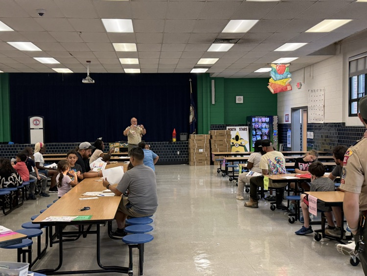An indoor view of a school cafeteria set up for Scout Night, featuring children and parents seated at long folding tables filling out papers, with a Scout leader in uniform standing near a stage in the background.