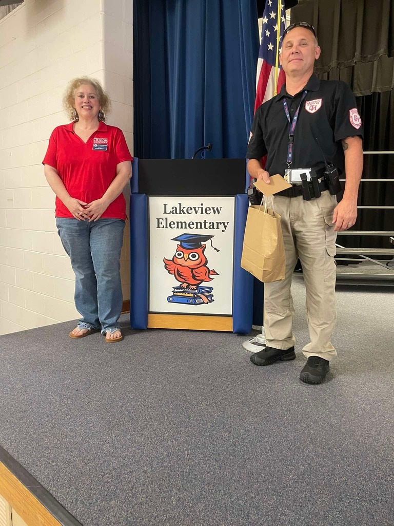 Two community partners pose beside the Lakeview Elementary podium following a visit to the school, with the school’s owl mascot displayed on the podium and the American flag in the background.