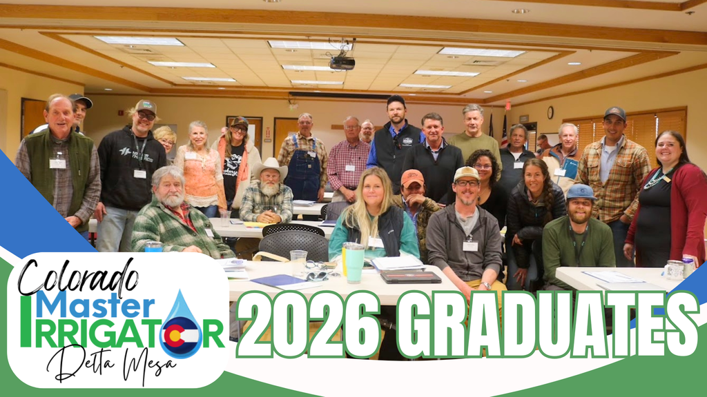 A group of approximately twenty people, the "Colorado Master Irrigator Delta Mesa 2026 Graduates," pose for a group photo in a brightly lit classroom or meeting room. They are a diverse group of men and women of various ages, dressed in casual attire like flannels, vests, hoodies, and baseball caps. Some are seated at white rectangular tables in the foreground, which hold notebooks, papers, and water bottles, while others stand behind them. In the lower-left corner, a green and white logo features the text "Colorado Master Irrigator Delta Mesa" alongside a stylized water droplet containing the Colorado state flag. Large, bold green letters across the bottom right of the image read "2026 GRADUATES.