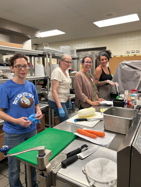 Food Service Team Preparing Meals Four women stand smiling in a commercial kitchen, surrounded by cutting boards, utensils, and fresh vegetables. They are mid-preparation, working collaboratively on meal components.