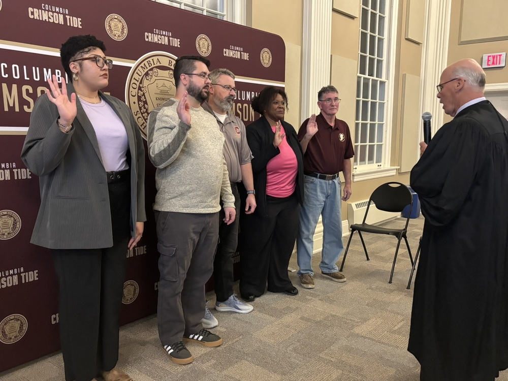 A group of five people stand in a row with their right hands raised as they participate in a swearing-in ceremony. They face an officiant dressed in judicial robes who holds a microphone. Behind the group is a maroon backdrop with the Columbia Crimson Tide logo. The setting appears to be an indoor hall with tall windows and beige walls.