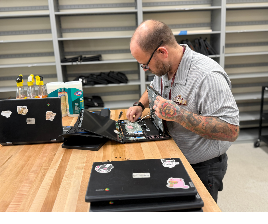 A technology staff member repairs an open laptop on a worktable, with additional laptops and computer equipment nearby.