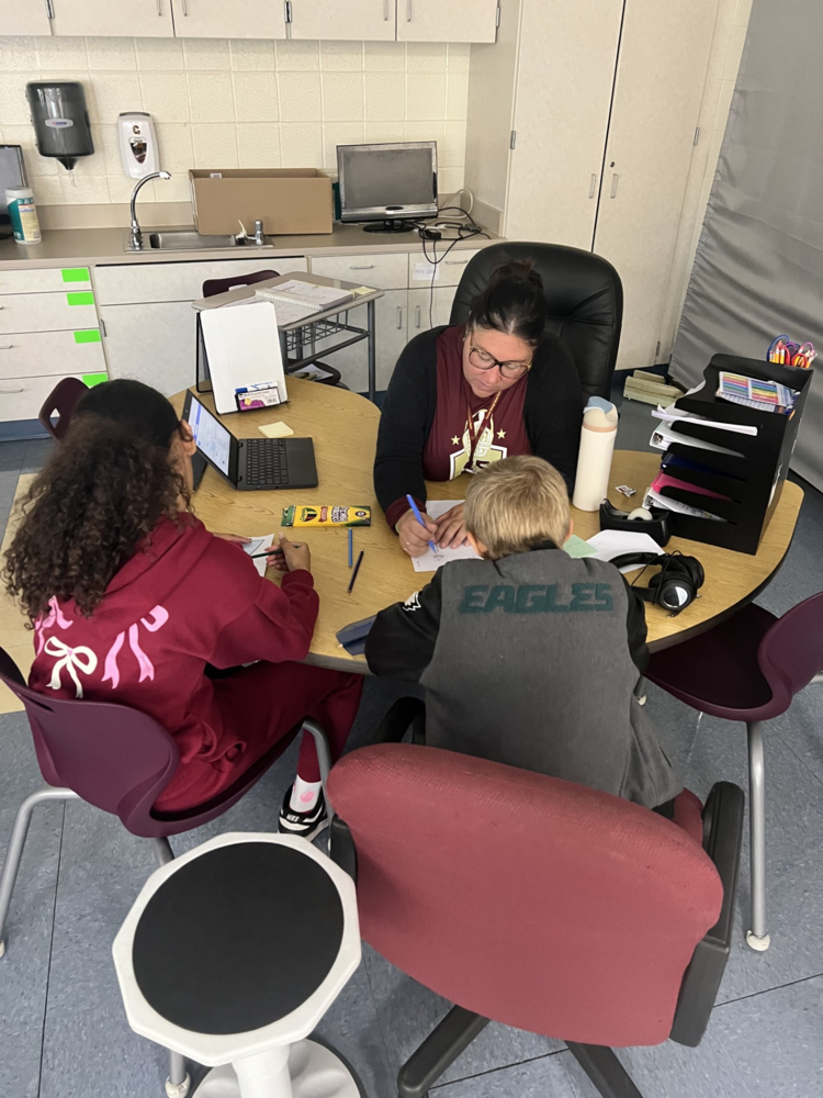 A teacher works one-on-one with two students at a round table, helping them complete a worksheet.