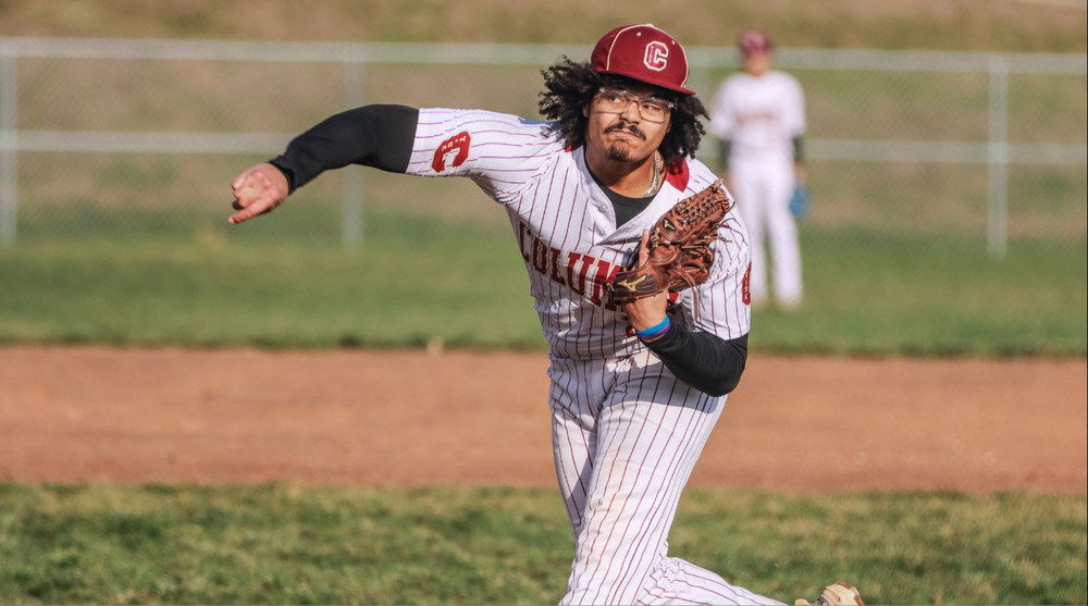 Columbia High School baseball player Peyton Gilbert delivers a pitch during a game while wearing a white pinstriped Columbia uniform with maroon lettering and a maroon cap featuring the school's "C" logo. Captured in mid-throw, Peyton extends his pitching arm forward while holding his glove close to his chest, showing focus and determination. His shoulder-length curly hair flows from beneath his cap, and he wears glasses as he concentrates on the batter. A teammate stands out of focus in the background, emphasizing the action on the pitcher's mound. The image highlights Peyton Gilbert's athleticism and intensity during live game competition.