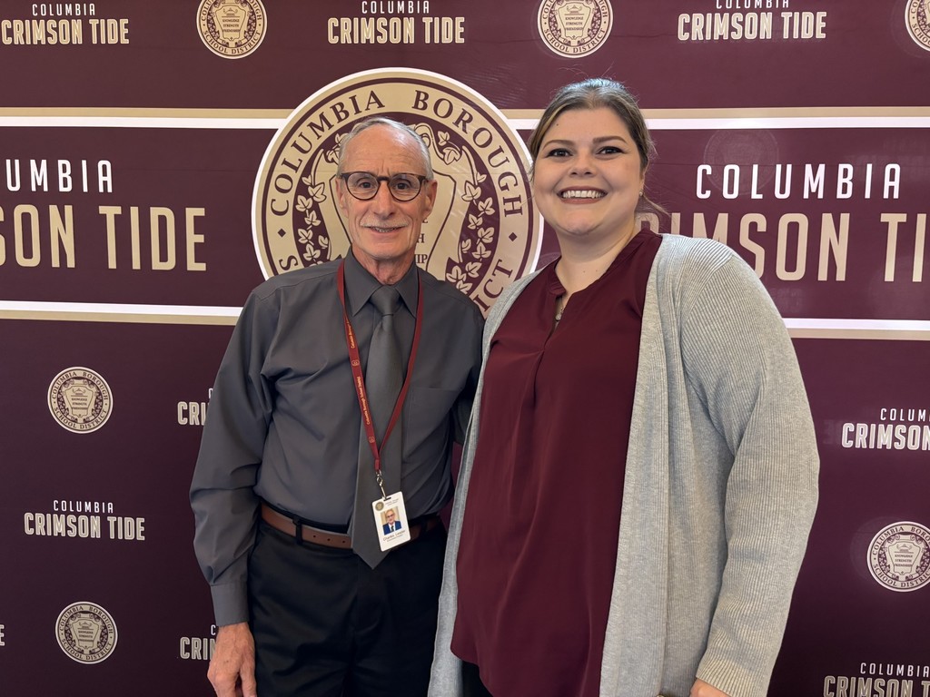 A smiling man and woman pose together in front of a Columbia Borough School District and “Columbia Crimson Tide” branded backdrop. The man, wearing glasses and a gray shirt with a matching tie and red lanyard, stands on the left. The woman, wearing a burgundy top and light gray cardigan, stands on the right, smiling broadly.