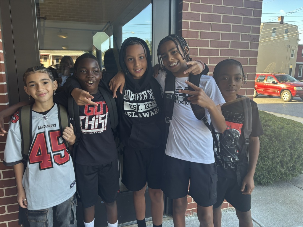 Group of friends posing by school entrance – Five middle school boys with backpacks smile and pose together outside by a brick wall.