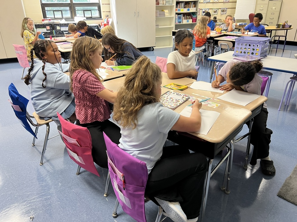 Elementary students sit in small groups at desks, drawing and writing on paper during a classroom activity.
