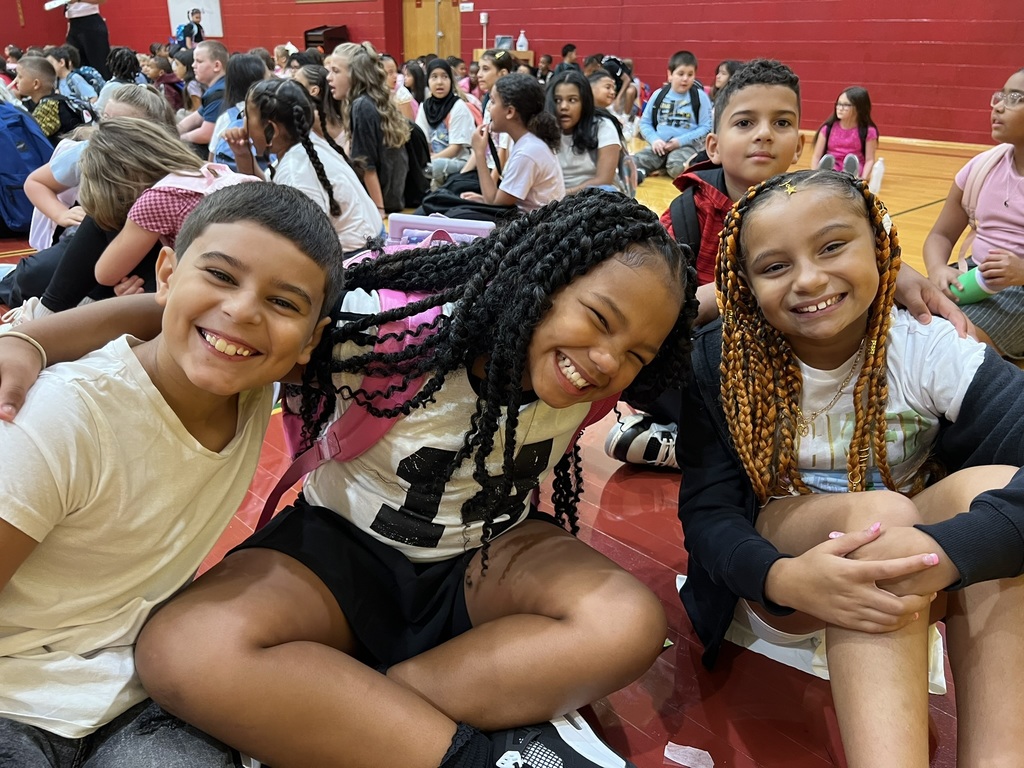 Students sitting together in the gym – Three elementary students sit on the gym floor with backpacks, smiling and leaning close for a photo with classmates around them.