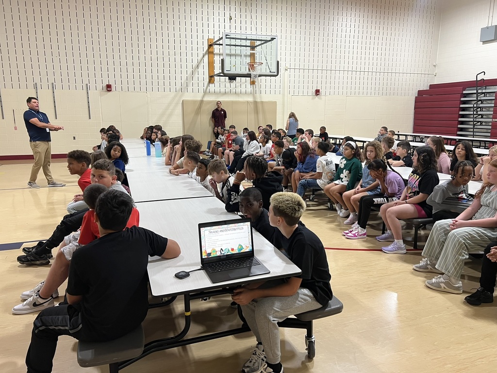 Students seated in a school gym – A group of middle school students sit at long cafeteria tables in the gym, listening to a staff member speak at the front of the room.