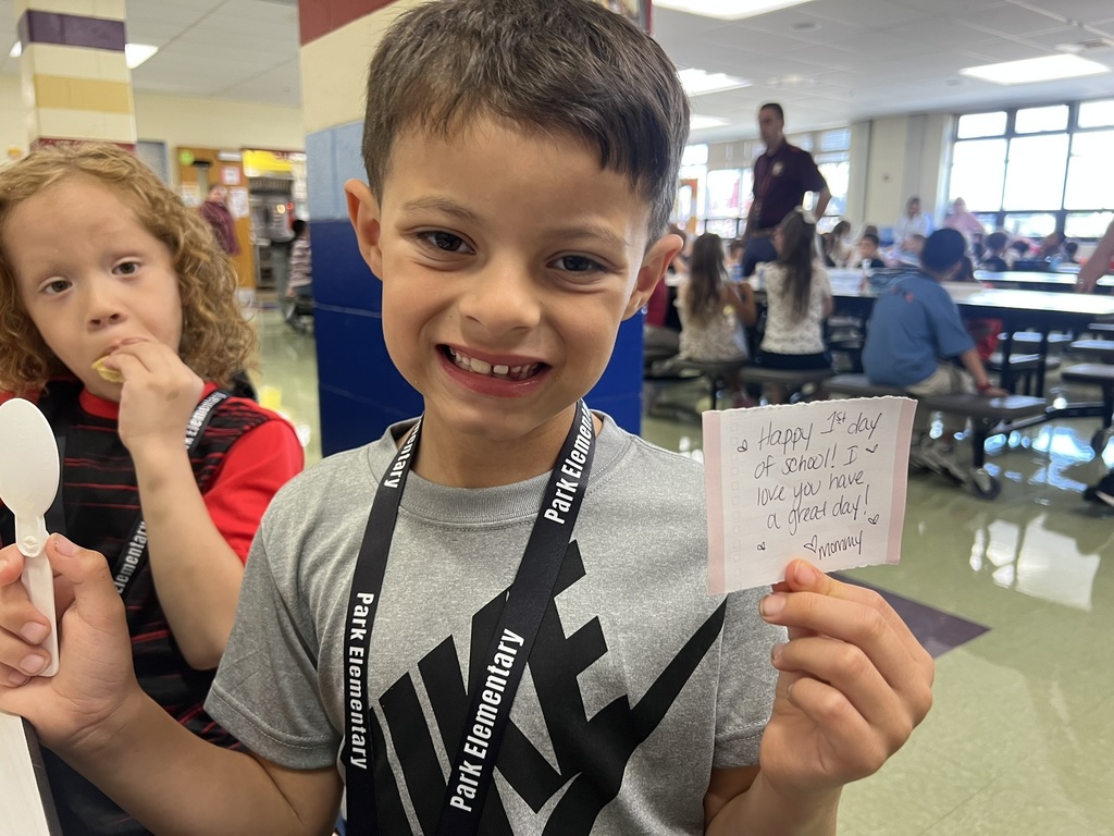 Student holding a first-day note – An elementary student smiles in the cafeteria while holding up a handwritten note from a parent wishing them a great first day.