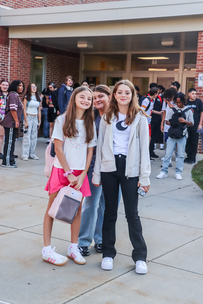 Three friends smiling outside school – Three girls stand smiling in front of the school entrance, surrounded by classmates arriving for the day.
