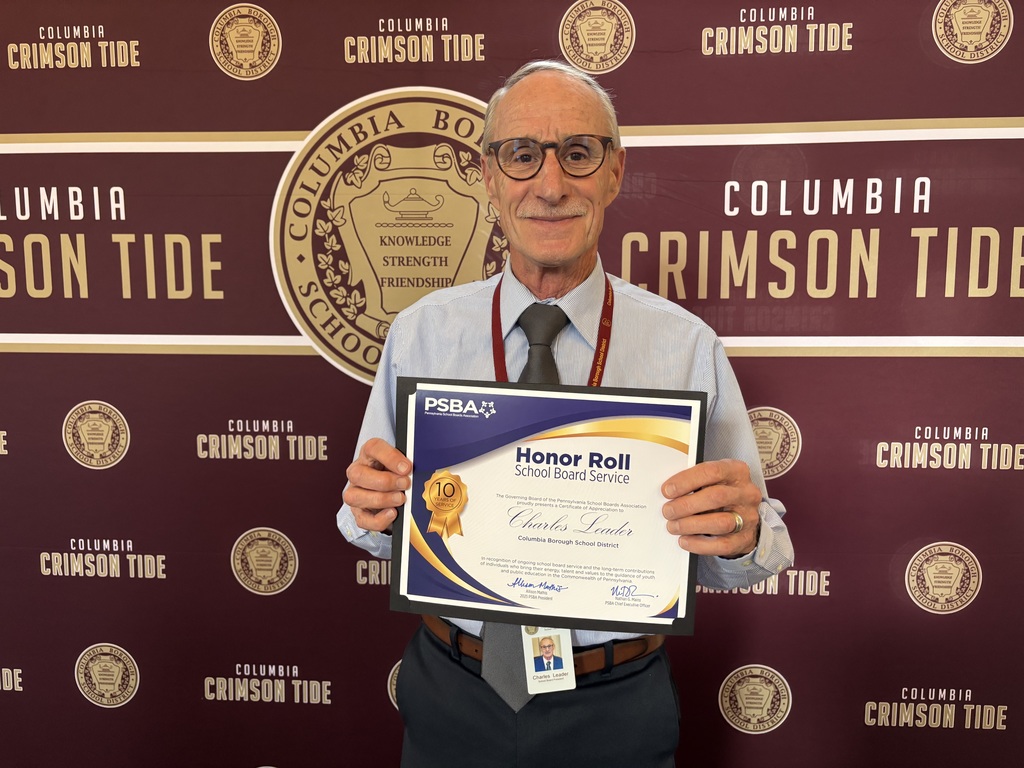 "Man wearing glasses, a light blue shirt, and a striped tie stands smiling in front of a Columbia Crimson Tide backdrop. He is holding a framed certificate from the Pennsylvania School Boards Association (PSBA) honoring him for 10 years of School Board Service. His name badge reads 'Charles Leader.'