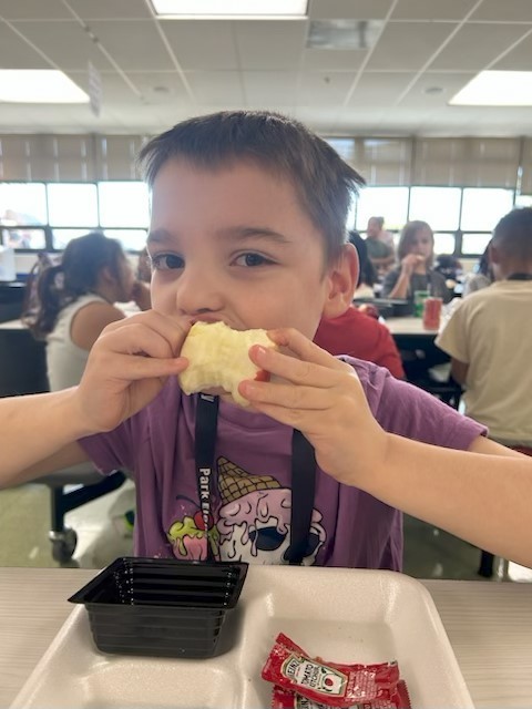 A young student in a purple shirt sits at a cafeteria table, holding a large apple with both hands and taking a big bite.