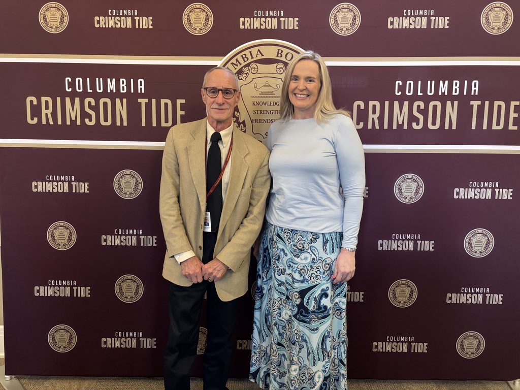 Charles Leader and Heather Lischner standing and smiling in front of a maroon backdrop featuring the words ‘Columbia Crimson Tide’ and the Columbia Borough School District seal. Mr. Leader, on the left is wearing glasses, a beige blazer, white shirt, and dark tie, while Ms. Lischner, on the right is wearing a light blue top and a long patterned skirt.