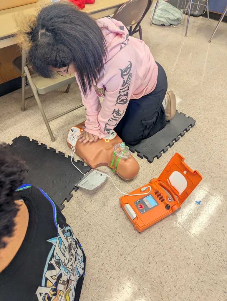 A student kneels on a mat performing chest compressions on a CPR training mannequin while an AED device is placed nearby on the floor.
